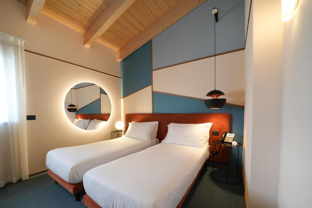 A hotel room with two beds, a round mirror, a bedside table with a lamp, and a phone.
