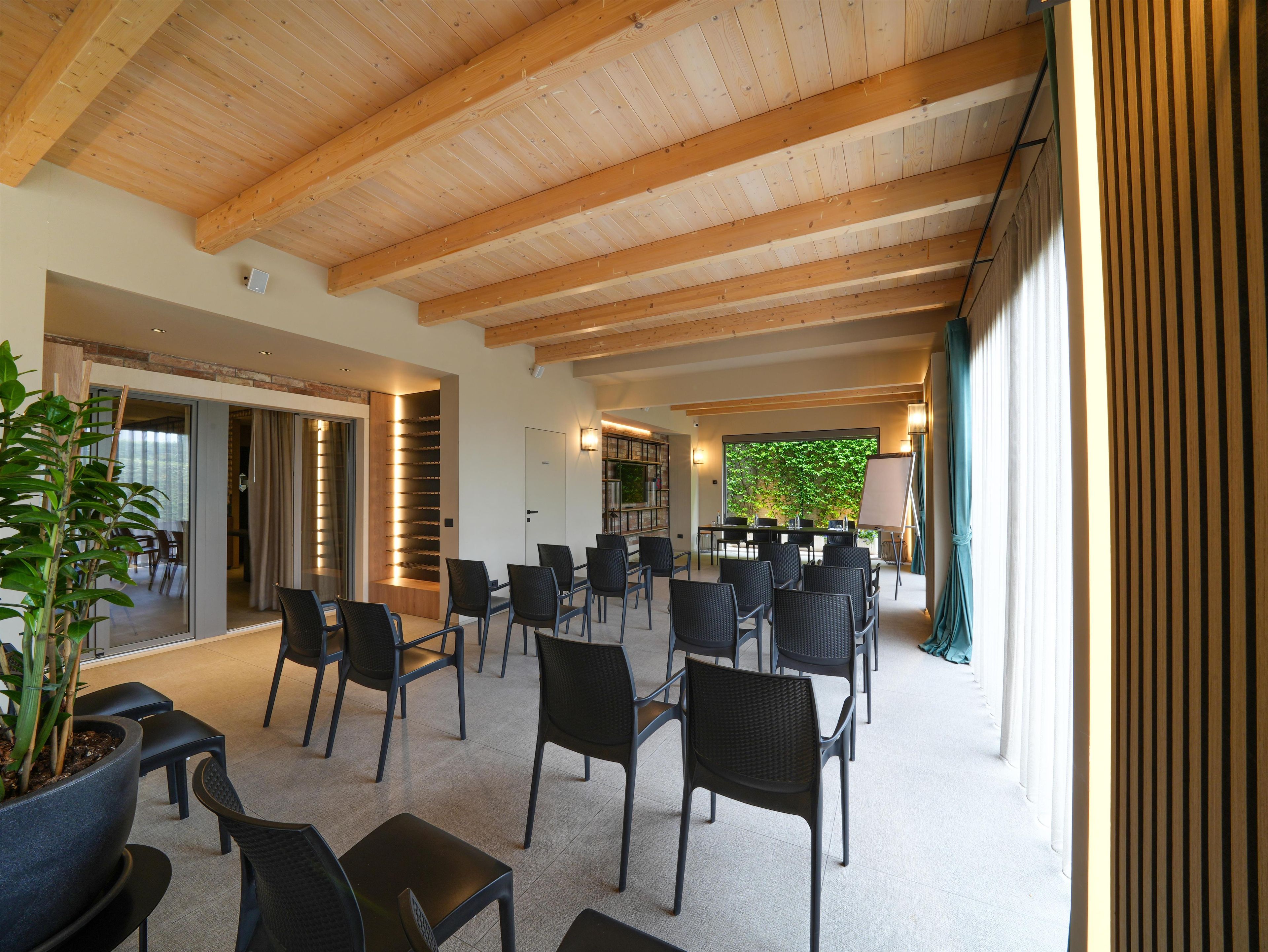 A spacious indoor area with wooden beams on the ceiling, a seating arrangement with chairs and tables, and a view of greenery outside.