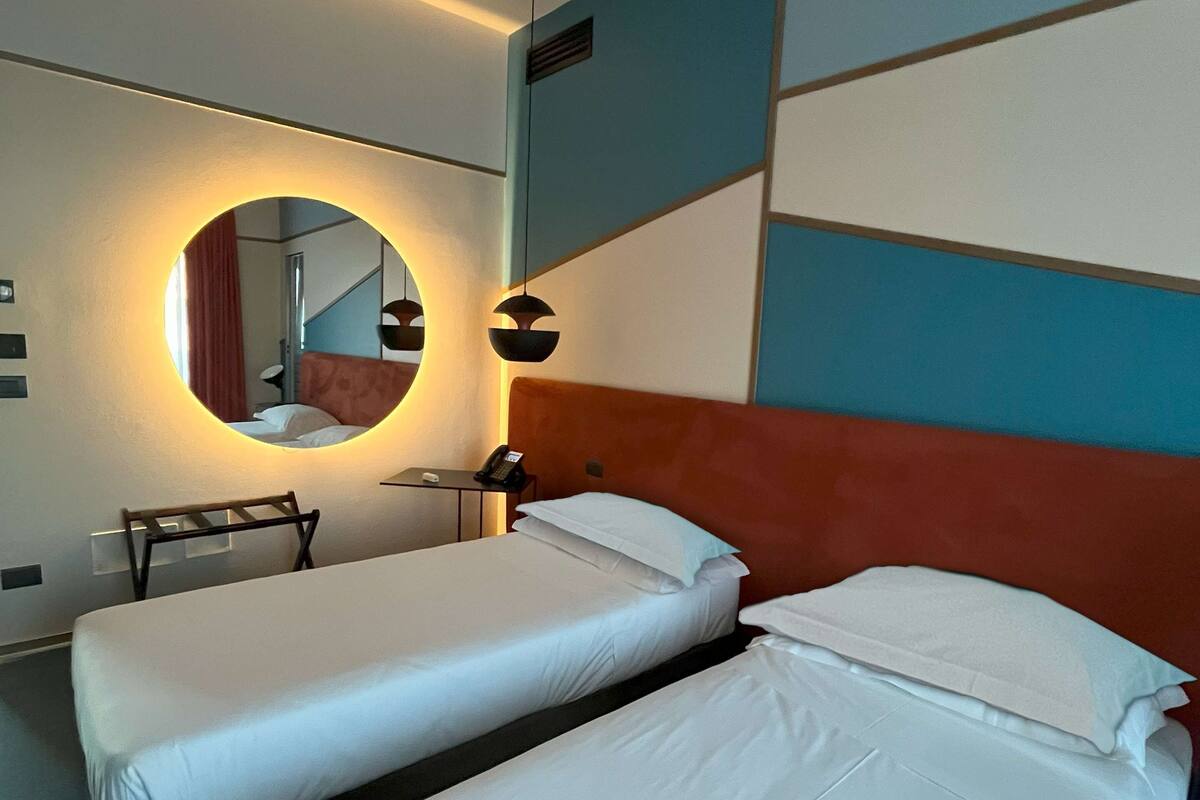 A hotel room with two single beds, a round mirror, a desk, and a chair.