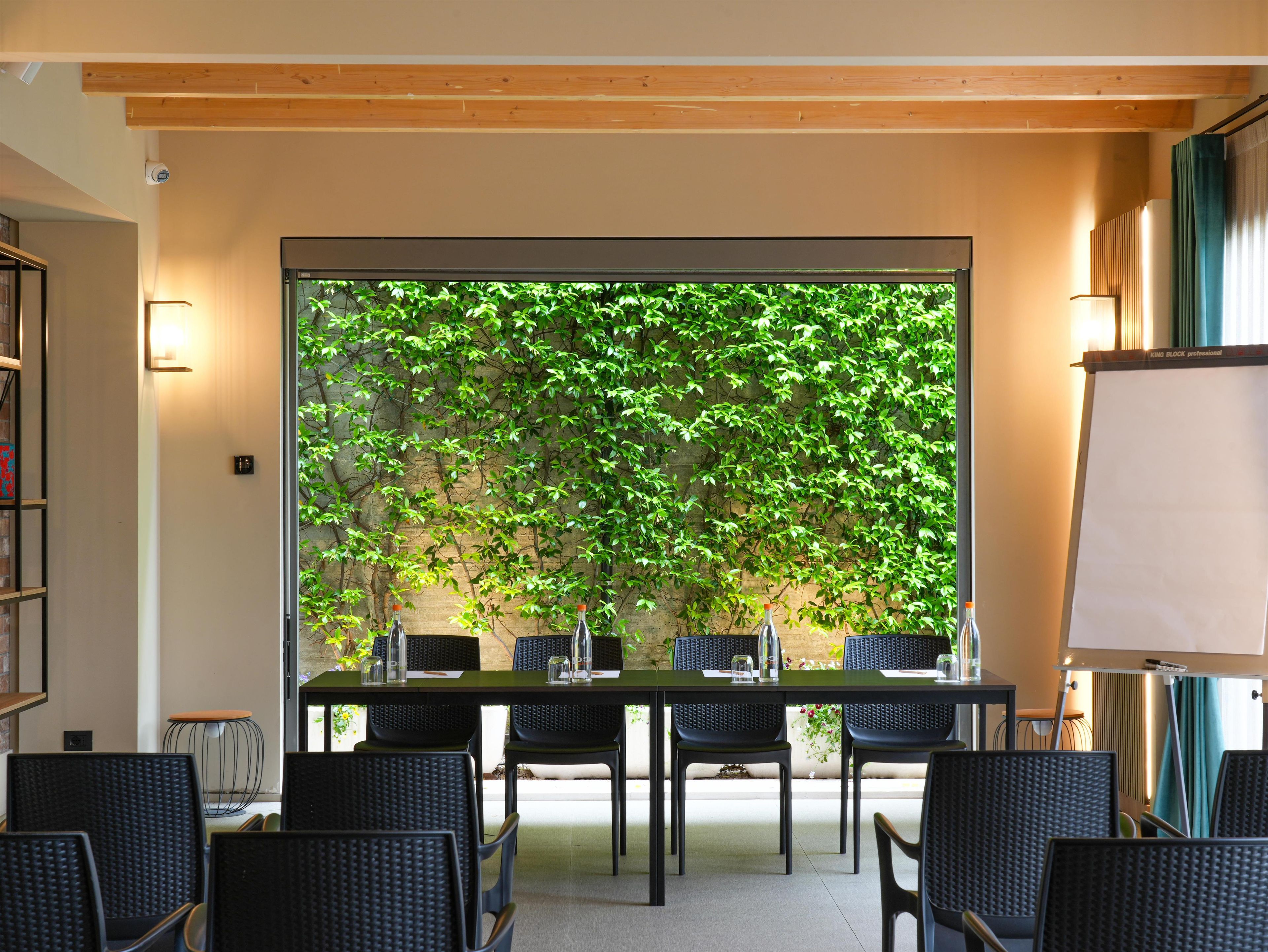 A conference room with a large table, chairs, a whiteboard, and a view of a green wall with climbing plants.