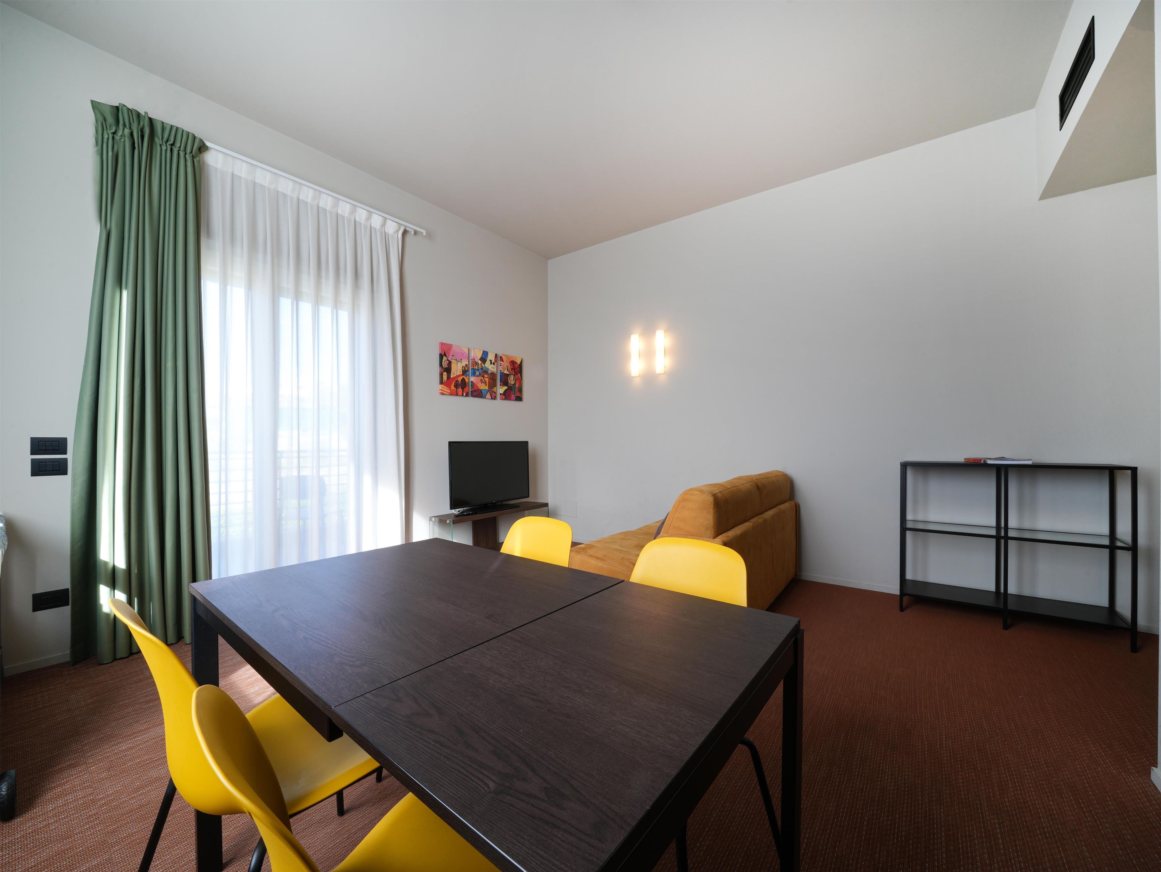 A modern room with a dark wooden table, yellow chairs, a brown sofa, a flat-screen TV, and a bookshelf.
