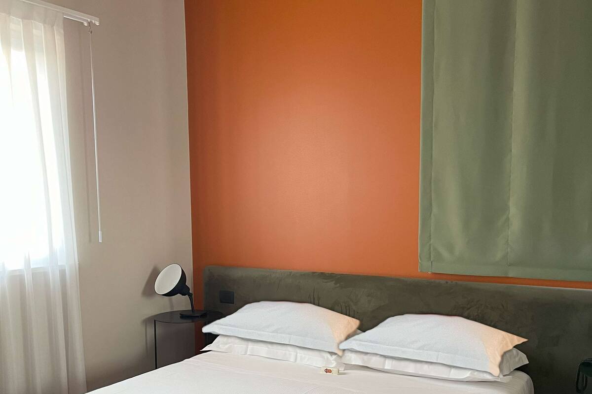 A neatly made bed with white linens in a room with an orange accent wall and a green curtain.