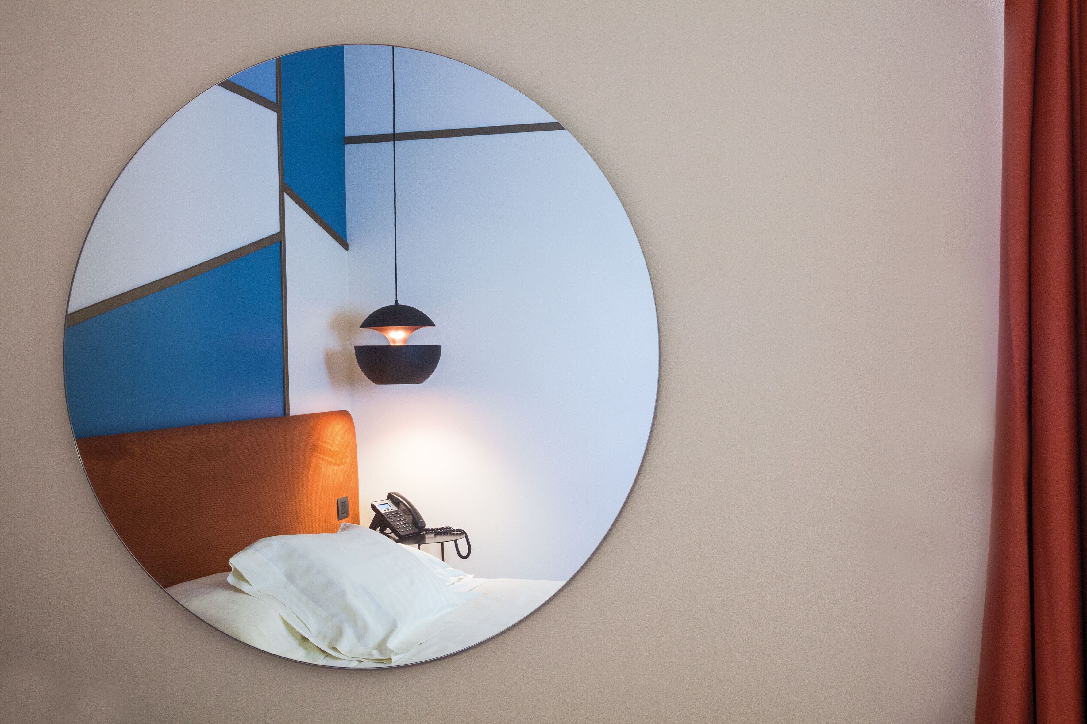 Uno specchio circolare che riflette un letto, un telefono, un lampadario e un motivo geometrico blu sul muro.