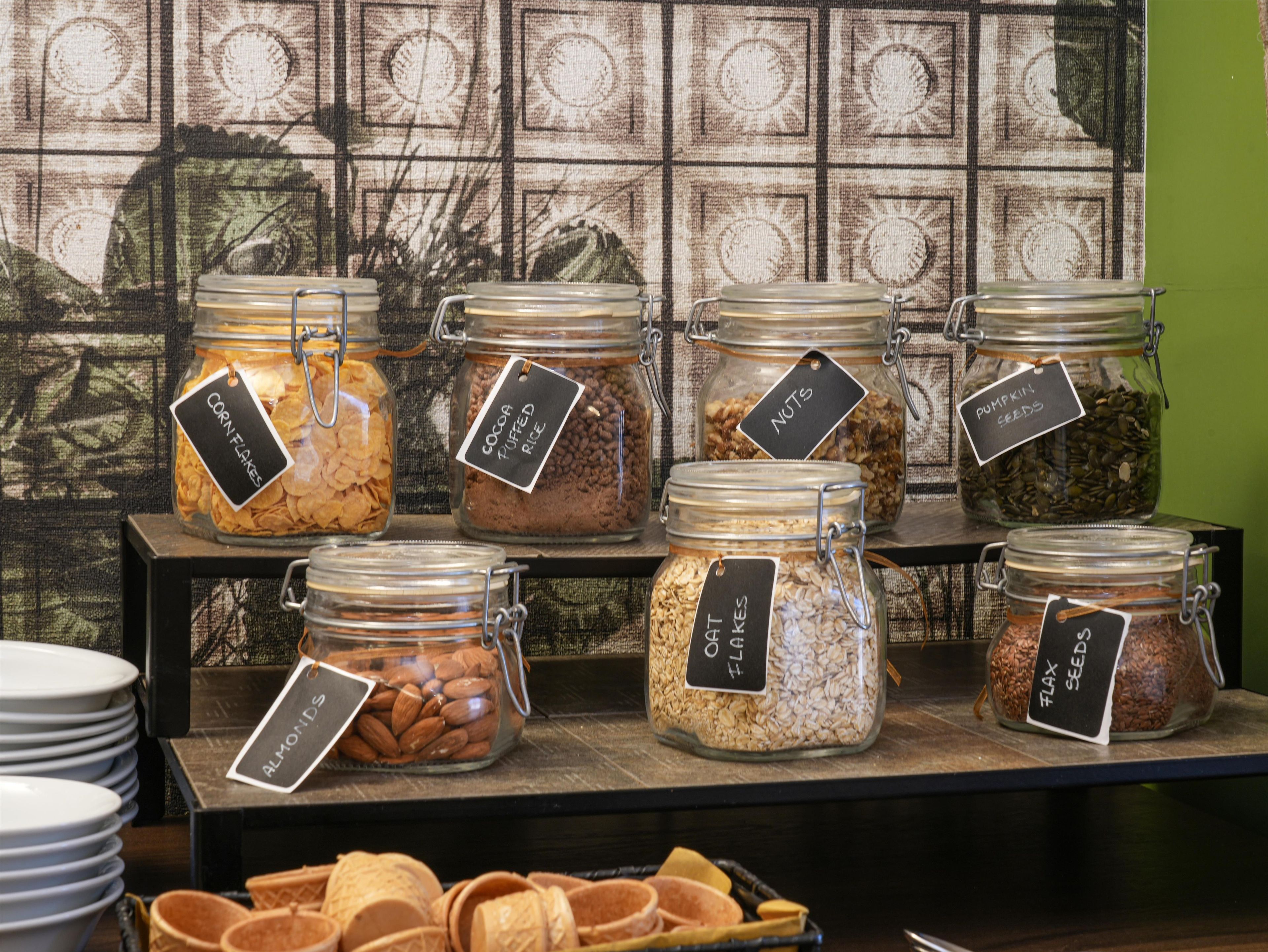 Une variété d’aliments sont présentés dans des bocaux en verre, chacun étant muni d’une étiquette, et l’ensemble est disposé sur une étagère.