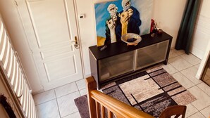 A room with a white door, a black console table with a painting, a patterned rug, and a staircase with a wooden handrail. - House close to the beach (Carnac)
