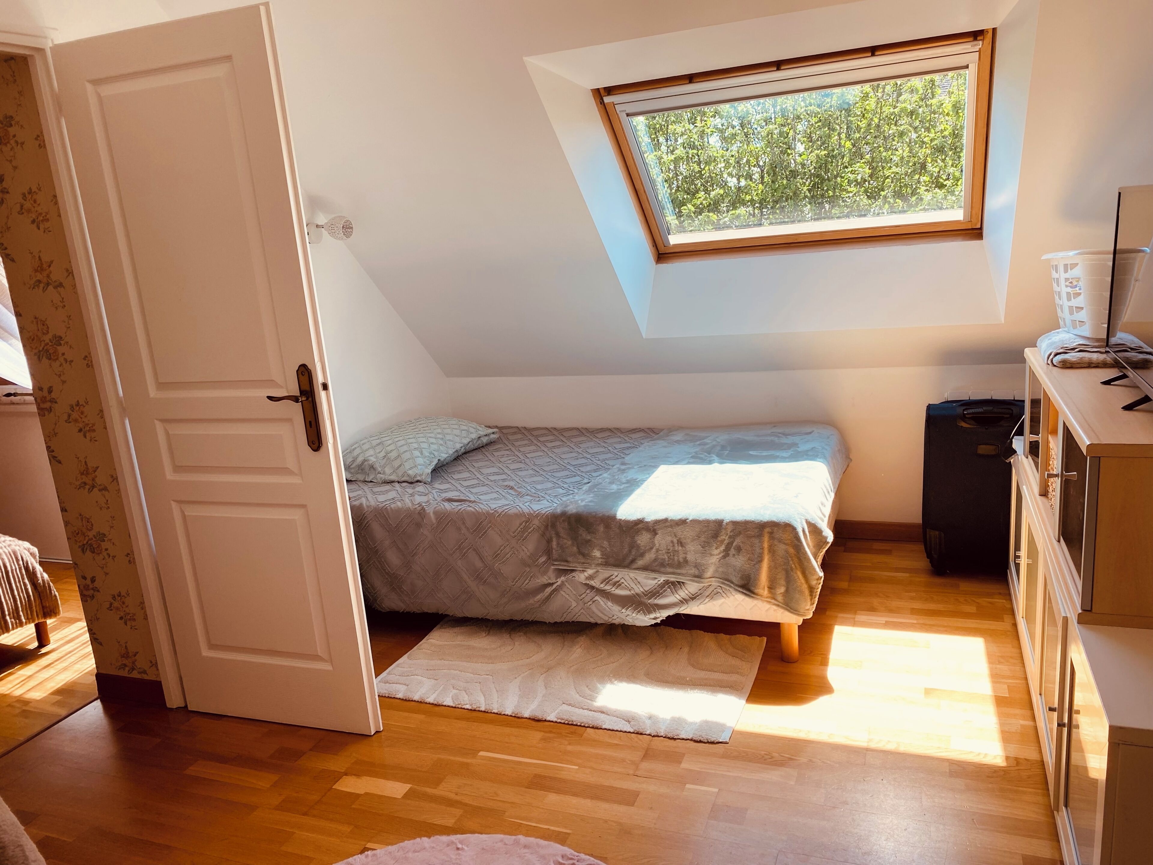 Ein kleines Schlafzimmer mit geneigter Decke, einem Fenster mit Blick auf Bäume, einem Bett mit gemustertem Überwurf, einer weißen Tür und einem Holzboden.