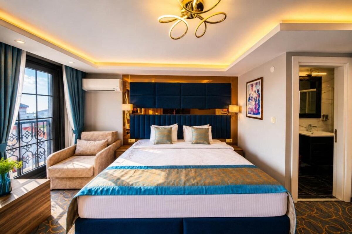 A hotel room with a large bed, a seating area with a chair, a wall-mounted air conditioner, and a view through a window.