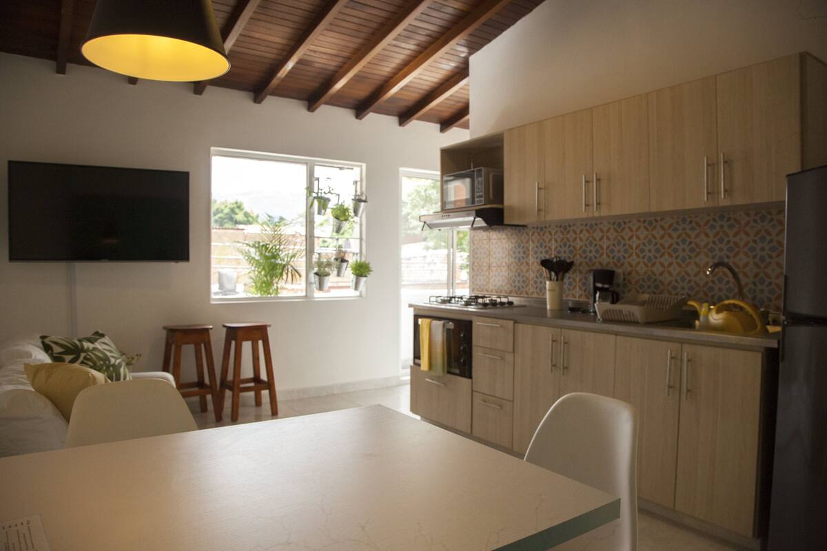 Cocina moderna con armarios de madera, microondas y estufa a gas. Hay dos taburetes y una mesa de comedor.