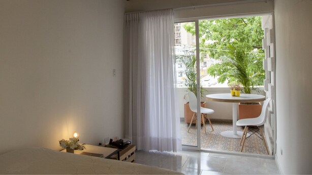 Habitación de hotel con cama, una mesita con lámpara, una silla y un balcón con mesa y sillas.