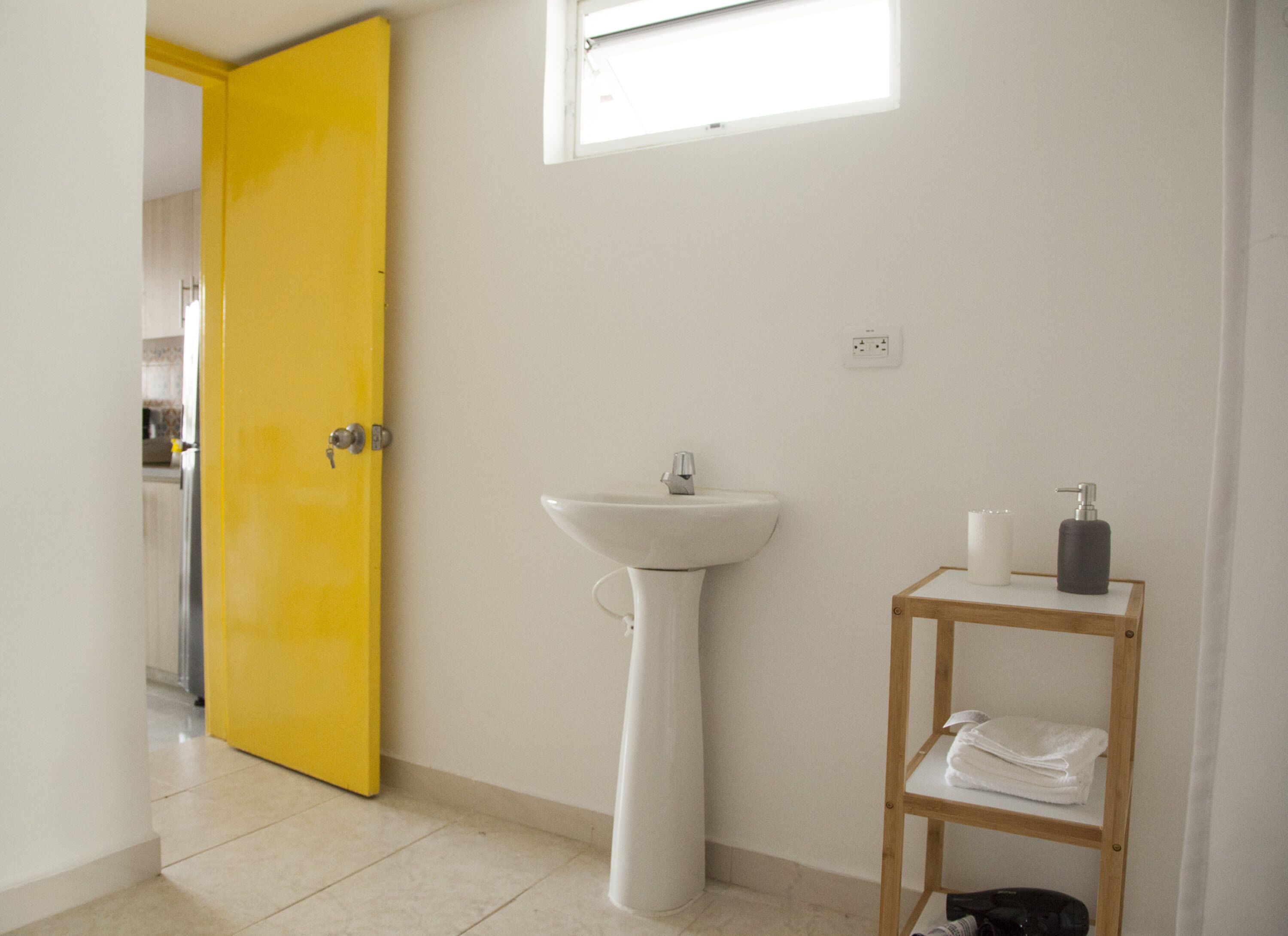 Un baño con una puerta amarilla, un lavabo blanco, un estante de madera con artículos de aseo y una ventana sobre el lavabo.