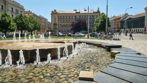 Une place publique avec une fontaine, des bancs en pierre et, en arrière-plan, des bâtiments historiques.