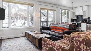 A modern living room with a leather sofa, a patterned armchair, and a coffee table. Large windows offer a view of the outdoors. - Outpost: King Ridge 4 - AC & Hot Tub! (Jackson)