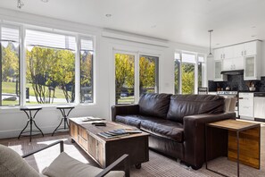 Una sala de estar moderna con un sofá de cuero, una mesa de centro de madera y vistas a un parque a través de amplios ventanales.