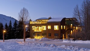 A modern building with a snow-covered roof and illuminated windows, surrounded by a snowy landscape and trees. - Outpost: King Ridge 4 - AC & Hot Tub! (Jackson)