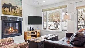 A living room with a fireplace, a television, a sofa, and a window with a view of trees. - Outpost: King Ridge 4 - AC & Hot Tub! (Jackson)