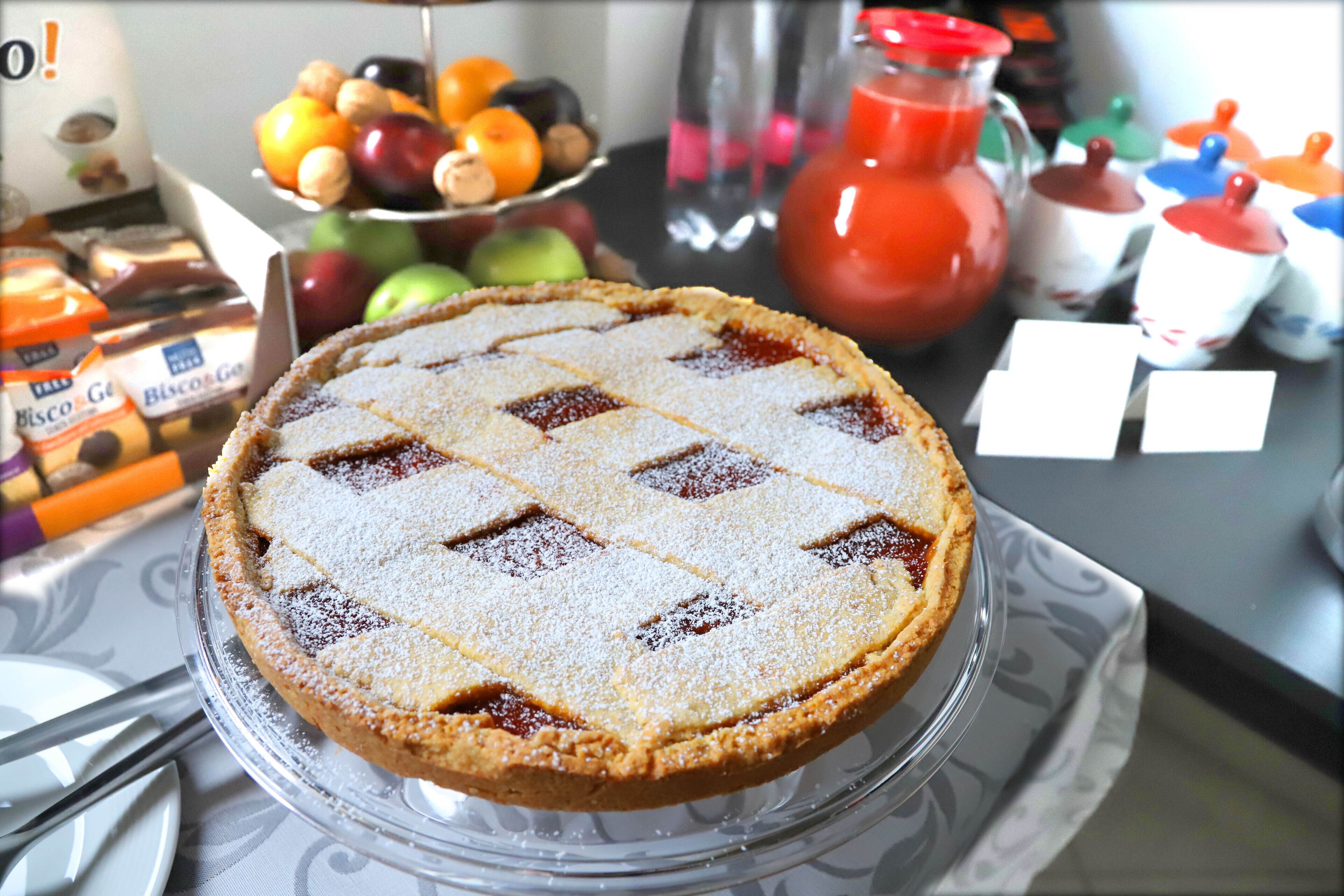 Una tarta sobre un pedestal de vidrio, con un frutero y condimentos al fondo.