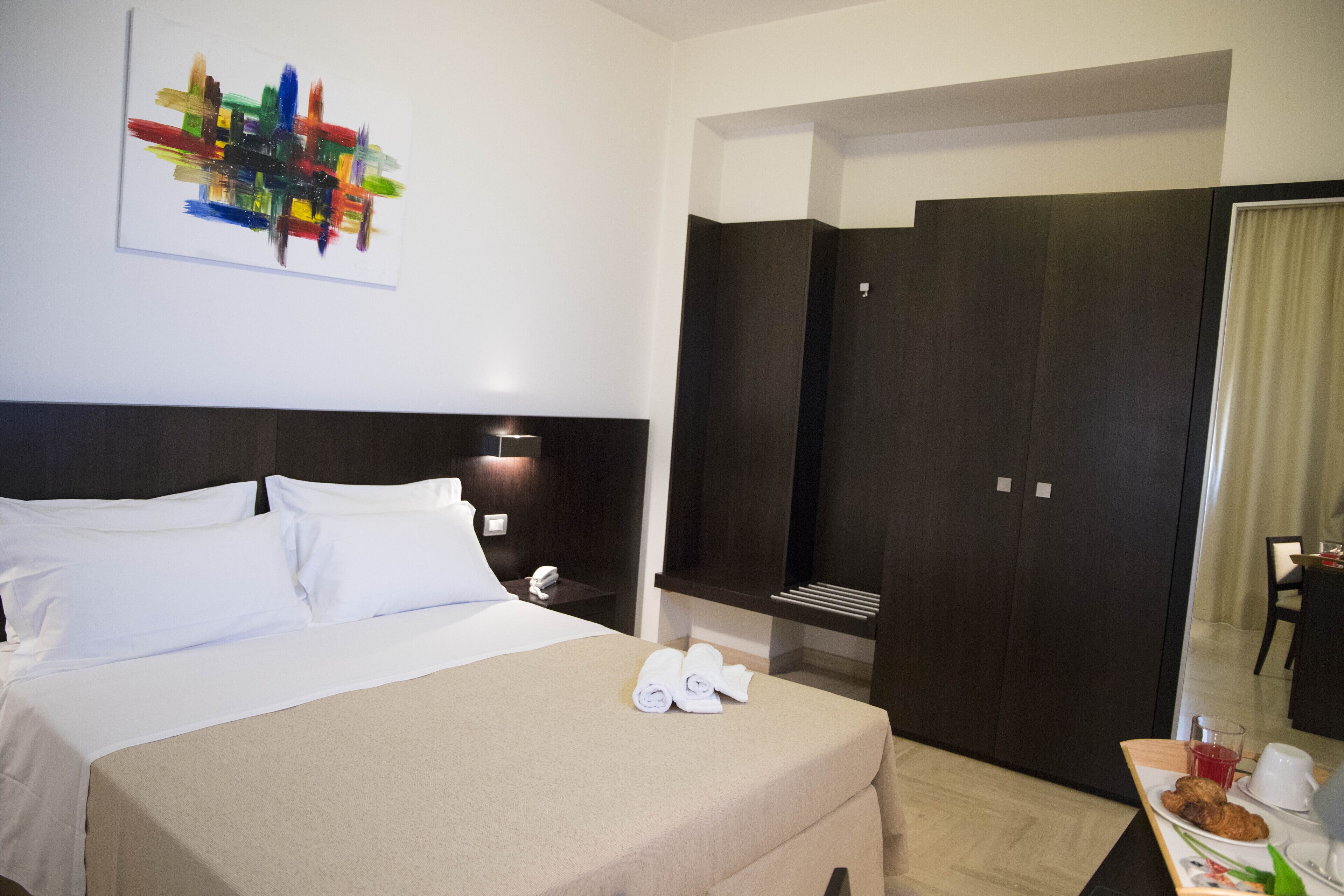 Ein Hotelzimmer mit Bett, einem Bild, einem Kleiderschrank, einem Schreibtisch mit Speisen und einem Spiegel.