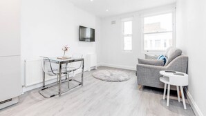 A modern living room with a grey sofa, a small white side table, a glass dining table with chairs, a round rug, and a mounted TV. - Flat 5 · Stunning 2-Bed Notting Hill Portobello Apartment (London)
