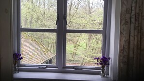 Una ventana con dos jarrones de flores moradas en el alféizar, con vistas a un jardín con árboles y un tejado de tejas.