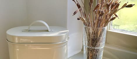 A white biscuit container with a handle and a glass vase with dried branches on a windowsill.