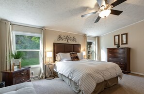 A bedroom with a large bed, a ceiling fan, a nightstand with a lamp, a dresser, and a window with curtains. - East Asheville - Views - Fire Pit - Parkway - Hiking - Pets - Wi-Fi - Large Home (Asheville)