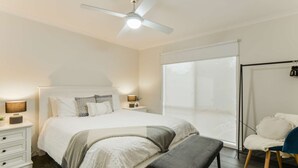 Une chambre à coucher comprenant un lit, des tables de chevet, un banc, une chaise, un ventilateur de plafond et une grande fenêtre.