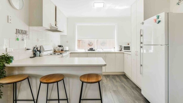 Une cuisine moderne avec des meubles blancs, un îlot central doté de tabourets, un réfrigérateur et une fenêtre avec des stores.