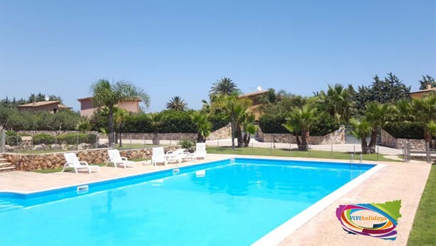 A swimming pool with lounge chairs, surrounded by greenery and buildings. - Viviholidays your holiday villa (Castellammare del Golfo)