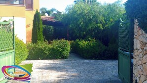 A stone pathway leading to a gated entrance surrounded by lush greenery and a building with a tiled roof. - Viviholidays your holiday villa (Castellammare del Golfo)