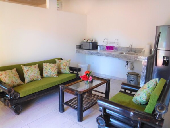 Un salón con un sofá verde, una mesa de centro de madera y un sillón verde. Se observa una zona de cocina al fondo con un refrigerador, un fregadero y un estante con utensilios de cocina.