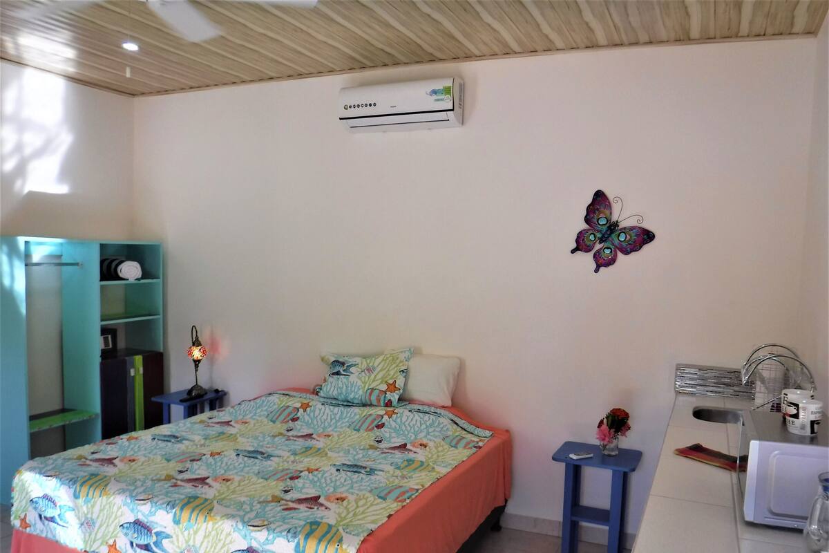 A bedroom with a bed, a desk, a chair, a shelf, and an air conditioning unit.