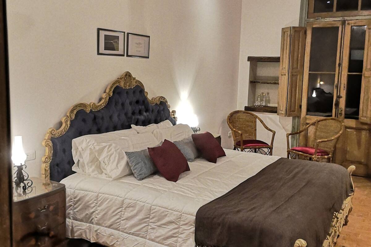 A bedroom with a large bed, wooden headboard, and a chandelier. There are two framed pictures on the wall and a window with wooden shutters.