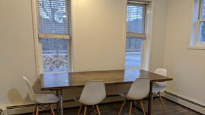 Ein Zimmer mit einem Holztisch und vier weißen Stühlen. Zwei Fenster mit Jalousien und ein Holzboden.