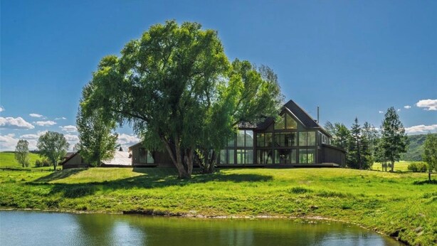 Une maison moderne dotée d’une grande façade vitrée, située au bord d’un plan d’eau et entourée d’une végétation luxuriante.