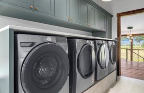Un espace buanderie équipé d’un lave-linge et d’un sèche-linge Whirlpool, avec des armoires turquoise au-dessus, et une vue sur une terrasse en bois par une porte ouverte.