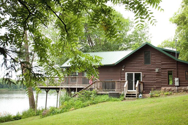 A rustic cabin with a deck overlooking a lake, surrounded by lush greenery. - Riverside Chalet, LLC (Muscoda)