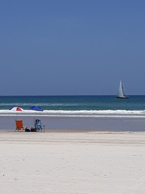 Une plage avec un voilier à l’horizon, un parasol rouge et bleu, et des chaises de plage.