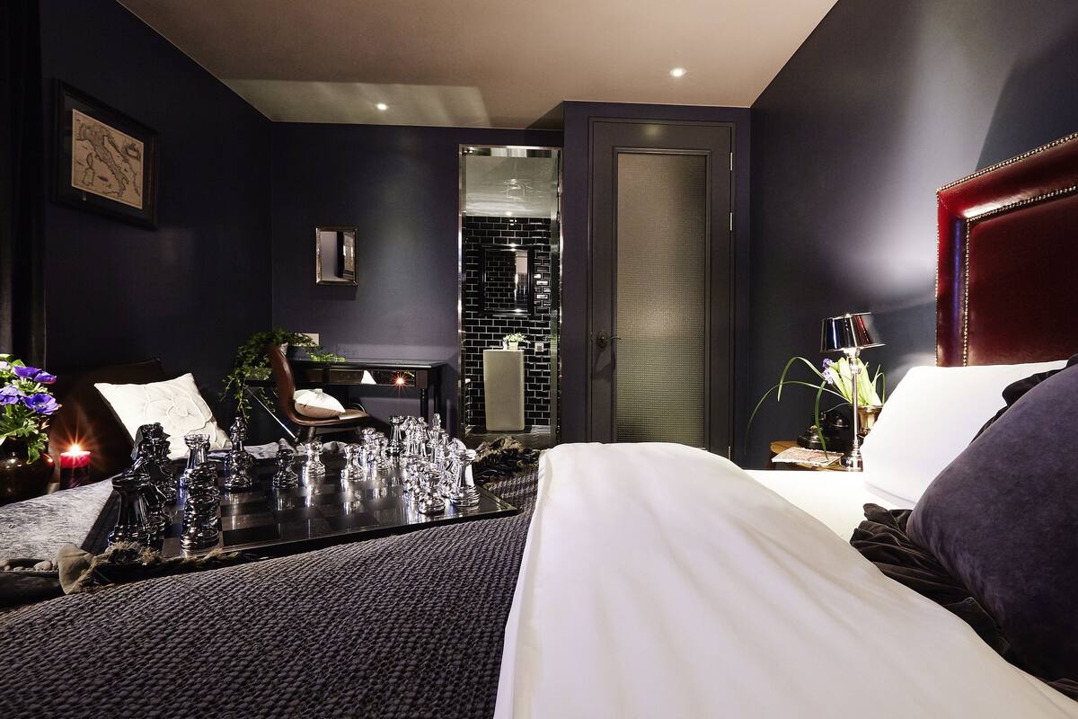 A modern hotel room with a large bed, a bedside table with decorative items, a chair, and a view into the bathroom.