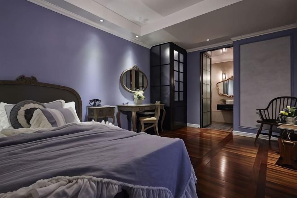 A bedroom with a large bed, a desk, and a chair. There is a mirror on the wall and a door leading to another room.