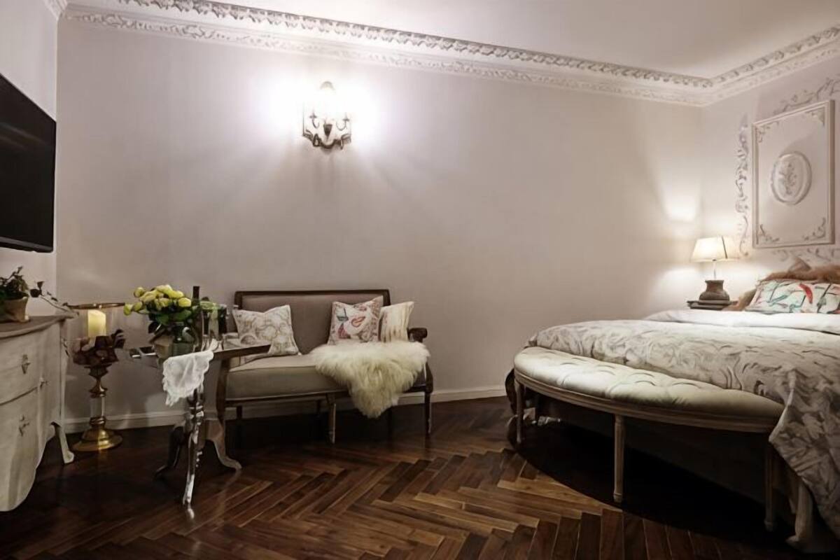 A bedroom with a bed, a bench, a bedside table with a lamp, and a wall-mounted light fixture.