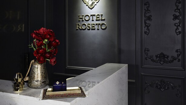 A reception desk with a vase of red roses, a gold-colored pitcher, and a small blue box. The wall behind features a sign with the text "HOTEL ROSETO". - Pocheon Hotel Roseto (Pocheon)