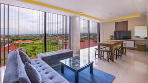 Uma sala de estar moderna com sofá, mesa de centro de vidro, mesa de jantar com cadeiras, televisão e vista para o horizonte urbano através de grandes janelas.
