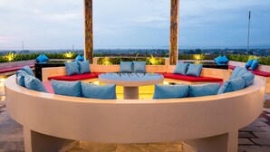 Uma área de estar ao ar livre circular com almofadas vermelhas e azuis, uma mesa central e vista para um horizonte urbano.