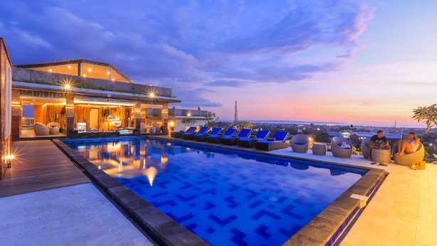 Piscina no terraço com espreguiçadeiras e área de bar, rodeada por um deck de madeira e com vista para a paisagem urbana ao entardecer.