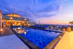 A rooftop pool with lounge chairs and a bar area, surrounded by a wooden deck and overlooking a cityscape at dusk. - Canggu Dream Village (Tibubeneng)