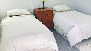 A simple room with two single beds, a wooden nightstand with a lamp, and a small drawer.