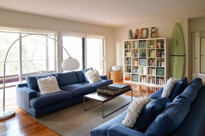 A modern living room with a blue sofa set, a coffee table, a bookshelf, and a surfboard. - Salt Bush - Great views nestled  in the moonahs (St Andrews Beach)