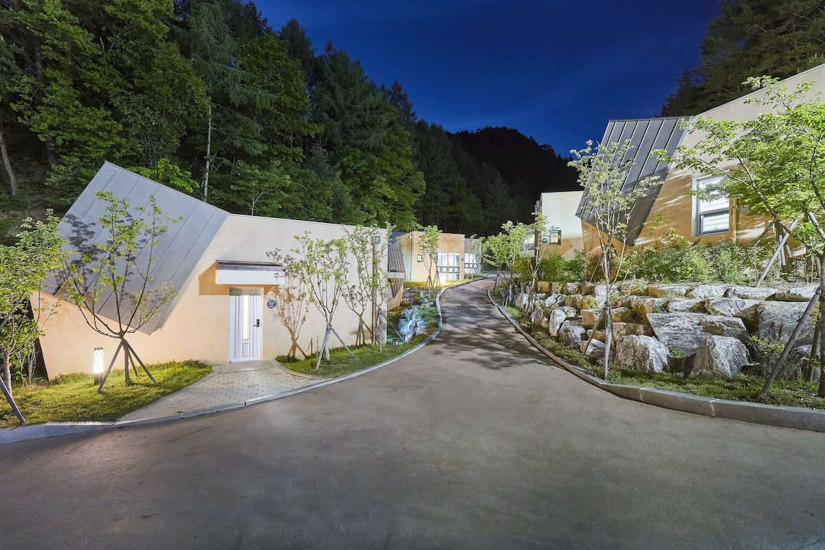 A residential street with modern houses, a paved road, and a forested hillside in the background.