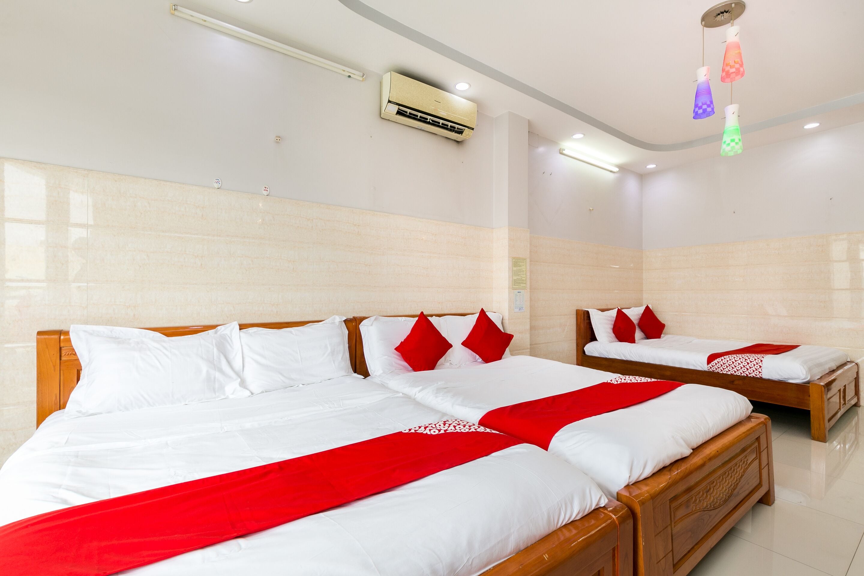 Une chambre d’hôtel avec trois lits, chacun doté de draps blancs et d’éléments décoratifs rouges, d’une tête de lit en bois et d’un sol carrelé.