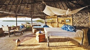 Una habitación con techo de paja, una cama, sillas y vista al mar.